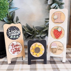 May include: Three wooden decorative signs. One sign has the text "Lucky Charm" and "Love is Sweet". Another sign has a sunflower. The third sign has a boot, strawberry, and ice cream cone with the text "Sweet Summer".