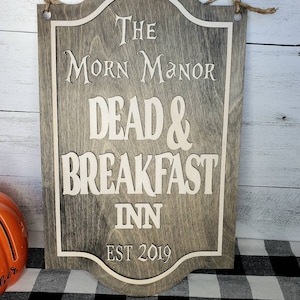 May include: A wooden sign with a gray background and white lettering. The sign reads "The Morn Manor Dead & Breakfast Inn Est 2019".