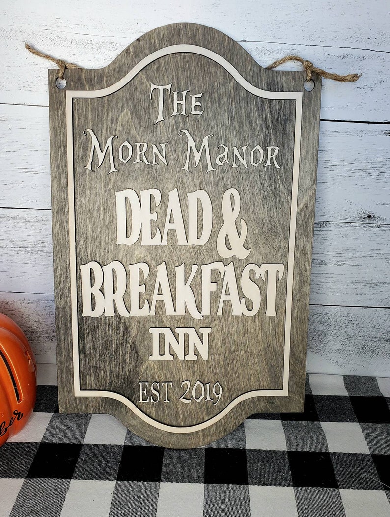 May include: A wooden sign with a gray background and white lettering. The sign reads "The Morn Manor Dead & Breakfast Inn Est 2019".