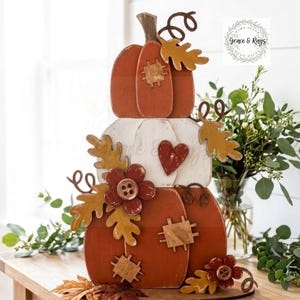 May include: A stacked wooden pumpkin decoration featuring three tiers in orange and white. The design includes painted patches, a red heart, wooden button flowers, and cutout oak leaves. A logo in the top right corner reads Grace & Rays.