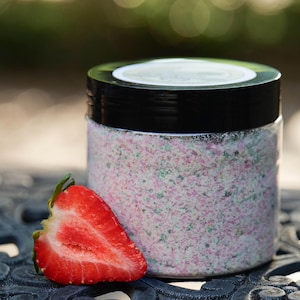 May include: A clear plastic jar filled with a granular mixture of pink and green particles, topped with a black screw-on lid. A fresh, halved strawberry with a bright red interior and green leafy stem is positioned directly in front of the jar.