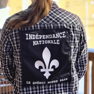 May include: A black and white plaid button-down shirt featuring a large rectangular black patch on the back. The patch displays the white text INDÉPENDANCE NATIONALE, a white fleur-de-lis symbol, and the text LE QUÉBEC NOTRE PAYS at the bottom.
