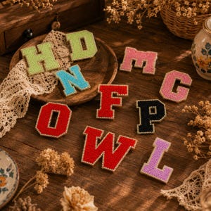 May include: A collection of varsity-style letter patches featuring a textured surface and a gold-toned beaded border. The set includes the letters H, D, N, M, G, O, F, P, W, and L in various colours including lime green, blue, pink, red, black, and lavender.