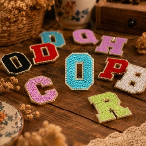 May include: A collection of varsity-style letter patches featuring a border of small white pearls. The letters are covered in sparkling rhinestones in various colors including light blue, red, black, pink, purple, white, and lime green.