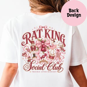 May include: A white t-shirt featuring a back graphic with the text "RAT KING Social Club" and "TROUBLE AFTER MIDNIGHT". The design includes a nutcracker, a ballerina, mice, cherries, ribbons, a drum, ballet slippers, and a crown in red and pink.