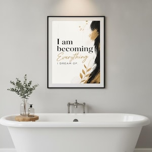 May include: A rectangular wall art print in a black frame featuring the text I am becoming Everything I DREAM OF. The design includes black and gold brushstroke accents and a gold leaf illustration on a white background, displayed above a bathtub.