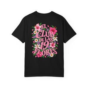 May include: A black short-sleeved t-shirt featuring a graphic on the back. The design displays pink text that reads EL CLUB DE LAS 19 FLORES, surrounded by a vibrant arrangement of pink and white hibiscus flowers, green leaves, and small purple blossoms.