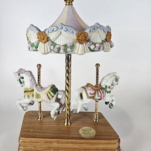 May include: A porcelain carousel music box featuring two white horses with pastel saddles and floral accents. The canopy is decorated with pink roses and blue trim, supported by a central gold-tone pole on a rectangular wooden base with a gold label.