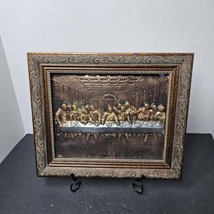 May include: A rectangular wall plaque depicting The Last Supper in a three-dimensional relief style. The scene features gold-toned figures seated at a long table, set within a dark bronze-colored frame with an ornate, embossed scrollwork border.