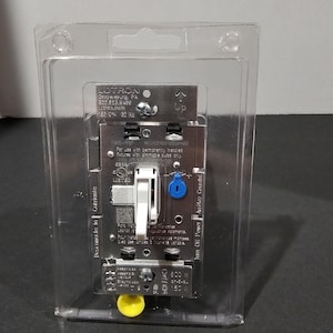 May include: A Lutron light dimmer switch with a white toggle lever, encased in clear plastic packaging. The metal faceplate features printed technical specifications, a blue adjustment dial, and a yellow wire connector at the bottom base.