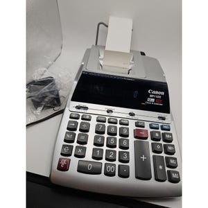 Puede incluir: Una calculadora impresora Canon MP11DX plateada con panel de pantalla negro, teclado numérico y un rollo de papel blanco cargado en la parte posterior. El dispositivo incluye teclas de función para cálculos de impuestos, margen y costes.
