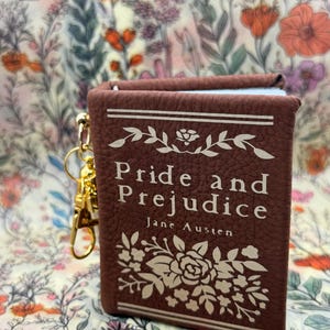 May include: A miniature book keychain with a textured brown cover featuring the title Pride and Prejudice by Jane Austen in white text. The cover is decorated with white floral illustrations and horizontal lines, and a gold metal lobster clasp is attached.