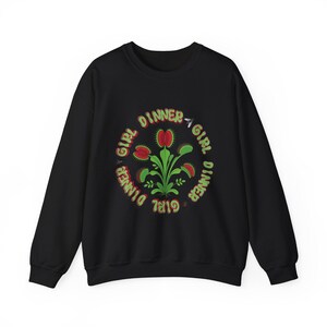 May include: A black long-sleeve sweatshirt featuring a central graphic of a green and red Venus flytrap plant. The text "GIRL DINNER" is repeated in a circular pattern around the plant in green and red letters, accompanied by small fly illustrations.