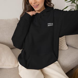 Puede incluir: Una sudadera negra de manga larga con capucha, cordón ajustable y bolsillo tipo canguro en la parte delantera. El texto AROID ADDICT está impreso en letras mayúsculas blancas en la parte superior izquierda del pecho, sobre un pantalón claro.