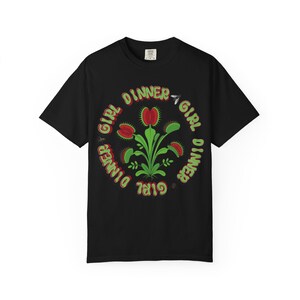 May include: A black short-sleeve t-shirt featuring a central graphic of a green and red Venus flytrap plant. The text "GIRL DINNER" is printed in a circular, wavy green font around the plant, accompanied by small illustrations of flies.