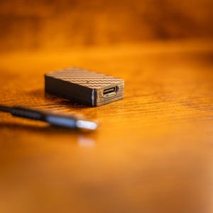 May include: A small, rectangular electronic adapter featuring a black carbon fibre pattern on its top surface and a visible USB-C port on its side, accompanied by a black charging cable with a matching USB-C connector head.