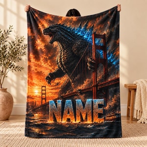 Op de afbeelding: Een rechthoekige fleece deken met een groot, donker, getextureerd monster dat achter de Golden Gate Bridge staat. De scène is geplaatst tegen een vurige oranje en blauwe zonsondergang boven water. Het woord NAME staat in grote letters gedrukt.