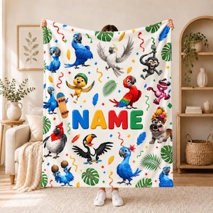 Könnte beinhalten: Eine weiße Fleecedecke mit einem bunten Muster aus verschiedenen Zeichentricktieren, darunter Papageien, ein Affe, ein Frosch und eine Bulldogge mit einem Obsthut. Das Wort NAME ist in großen, mehrfarbigen Blockbuchstaben in der Mitte gedruckt.