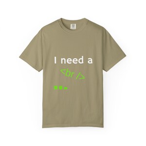 May include: A khaki-colored short-sleeve t-shirt featuring the white text "I need a" followed by the lime green HTML tag "<br />" and three small lime green square icons arranged horizontally on the front of the garment.