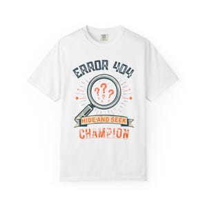 May include: A white short-sleeve t-shirt featuring a graphic print with the text ERROR 404, a magnifying glass containing three orange question marks, and a banner with the text HIDE AND SEEK CHAMPION in orange and dark blue ink colours.