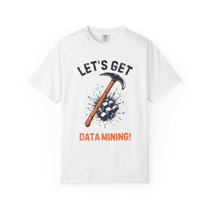 May include: A white short-sleeve t-shirt featuring a graphic of an orange-handled pickaxe striking a cluster of blue-tinted digital cubes. The text "LET'S GET" appears in black above the graphic, and "DATA MINING!" is printed in orange below.