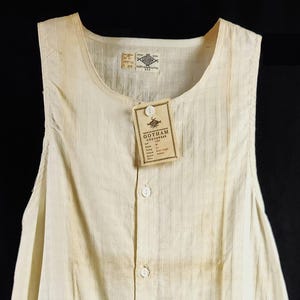 May include: Cream-colored, sleeveless one-piece garment with vertical stripes and a button-down front. A small tag is attached near the neckline. The garment appears to be made of a lightweight fabric.