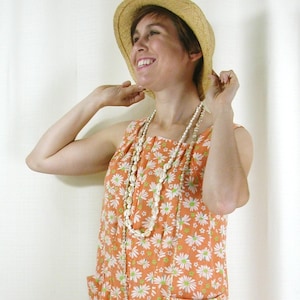 May include: A woman wearing a straw hat and a sleeveless dress with a white daisy print on an orange background. She is wearing a long white beaded necklace.