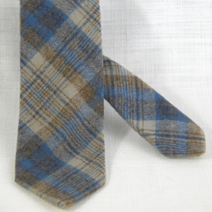 May include: A patterned necktie in blue, beige, and brown. The tie has a classic plaid design, with a diagonal arrangement. The fabric looks to be woven. The tie is shown against a white backdrop.