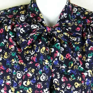 Può includere: Una blusa blu navy con un grande fiocco al collo. La blusa ha una stampa astratta colorata con accenti bianchi, gialli, rosa e verdi.