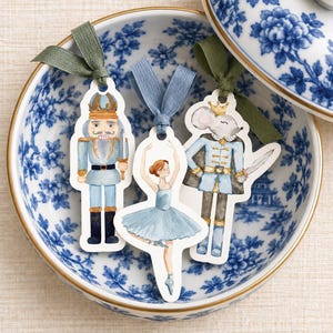 Peut inclure: Trois marque-pages en papier découpé présentant des illustrations à l'aquarelle d'un casse-noisette, d'une ballerine et d'un roi souris. Chaque marque-page est surmonté d'une boucle en ruban de tissu vert sauge ou bleu cendré.