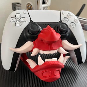 Puede incluir: Un mando de videojuegos blanco y negro con un accesorio de máscara impreso en 3D de color rojo y crema. La máscara cubre el centro del mando y muestra un rostro estilizado con colmillos prominentes, una nariz y una pieza para la barbilla.