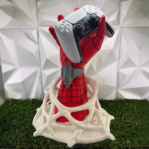 Puede incluir: Una escultura de mano con patrón rojo y negro que emerge de una base de telaraña blanca, sosteniendo un mando de videojuego blanco y negro. La mano presenta un accesorio de muñeca gris y está colocada en posición vertical con textura.