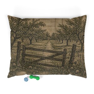 May include: A rectangular throw cushion featuring a woven tapestry design of a wooden gate leading into an orchard with trees and a small building in the distance. The colour palette consists of shades of brown, tan, and dark green. A small blue ball and a green dumbbell-shaped toy are placed in front of the cushion.