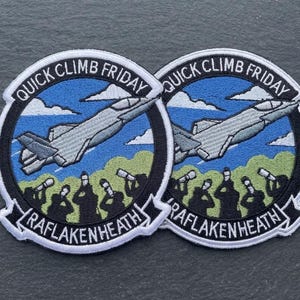 May include: Two circular embroidered patches featuring a grey fighter jet flying over a green landscape and blue sky with white clouds. The black border contains the white text QUICK CLIMB FRIDAY at the top and RAFLAKENHEATH at the bottom.
