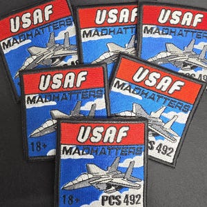 May include: A collection of square embroidered patches featuring a red top section with the white text USAF and a blue bottom section with the white text MADHATTERS. Each patch depicts a grey fighter jet flying over white clouds, with some patches displaying the text 18+ and PCS 492.