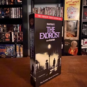 May include: A black VHS tape box for The Exorcist, featuring a silhouette of a person standing under a streetlamp at night. The title is printed in large purple letters, with the text The Version You've Never Seen and The Scariest Movie Of All Time.