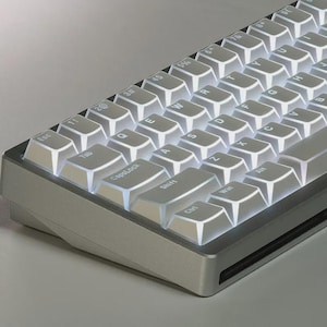 Könnte beinhalten: Eine mechanische Tastatur mit einem silbernen Metallrahmen und weißen Tastenkappen mit schwarzer Beschriftung. Die Tasten sind mit hellem weißem Licht hinterleuchtet, das die Zwischenräume beleuchtet. Das Layout ist QWERTY.