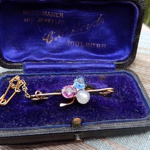 May include: A gold bar brooch featuring three faceted stones in pink, blue, and white, attached to a small safety pin by a delicate gold chain. The brooch is presented inside a black rectangular jewellery box with a deep blue velvet interior.