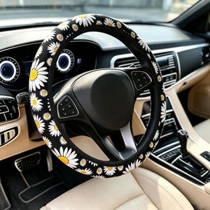 May include: A black steering wheel cover featuring a repeating pattern of white daisy flowers with yellow centers. The cover is fitted onto a black steering wheel inside a vehicle with a light-colored interior, dashboard, and center console controls.