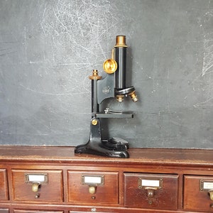 Könnte beinhalten: Ein antikes schwarzes Mikroskop mit Messingakzenten steht auf einem Holzschrank mit mehreren Schubladen. Der Schrank hat Messinggriffe und Etiketten an jeder Schublade.