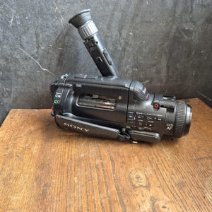 May include: A black Sony camcorder with a viewfinder and a handle. The camcorder has various buttons and dials, including "CCD AF" and "SONY" branding. It rests on a wooden surface against a dark background.