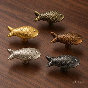 Könnte beinhalten: Ein Set aus fünf fischförmigen Schrankknöpfen in verschiedenen metallischen Ausführungen wie Gold, Bronze, Silber und Dunkelgrau. Jeder Knopf hat ein strukturiertes Rautenmuster als Schuppen und ein kleines kreisförmiges Auge. Der Text VllnCo ist sichtbar.