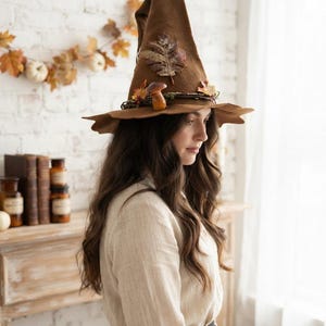 May include: A brown felt witch hat featuring a tall, pointed crown and a wide, uneven brim. The hat is decorated with a dark vine wrapped around the base, adorned with a single brown mushroom and several colourful autumn leaves.
