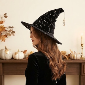 May include: A black velvet witch hat featuring a pointed crown and a wide brim, decorated with a gold star pattern. A delicate silver-toned chain with small star-shaped charms hangs from the tip of the hat, adding a subtle metallic accent.