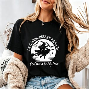 May include: A black short-sleeved t-shirt featuring a white graphic of a witch flying on a broomstick against a full moon. The text reads ON A DARK DESERT HIGHWAY and Cool Wind In My Hair, surrounded by small bats and stars in a circular design.