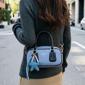 Puede incluir: Un bolso de mano de mezclilla azul claro con asas y bordes de cuero negro, que cuenta con un cierre de cremallera plateado y un dije colgante en forma de estrella de mar azul con cuentas blancas en el lateral.