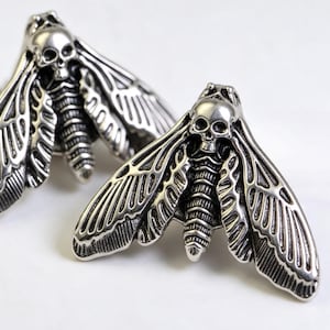 May include: Two silver-toned metal pins shaped like moths, each featuring a detailed skull design on the thorax. The wings display intricate, raised line patterns and textures, with a polished finish that highlights the anatomical details of the insect.