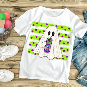 Może przedstawiać: Biały t-shirt z grafiką przedstawiającą białego ducha trzymającego fioletową puszkę napoju energetycznego. Tło grafiki składa się z poziomych, limonkowych pociągnięć pędzla z fioletowymi kropkami. Na puszce znajduje się napis Alani.