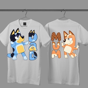 May include: Two white short-sleeve t-shirts hanging on hangers. The left shirt features a blue cartoon dog and the text DAD. The right shirt features an orange cartoon dog and the text MUM. Both shirts have a crew neckline and a straight hem.