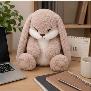 May include: A plush stuffed animal in the shape of a rabbit with long, floppy ears. The rabbit has light brown, textured fur, a white face and chest, and a small brown nose. The word BANA is embroidered on the bottom of the left foot.